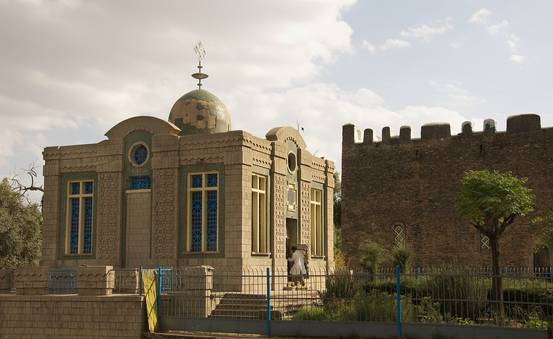 A domed stone chapel with tall blue windows behind a low fence at Aksum, with an old battlemented church beside it