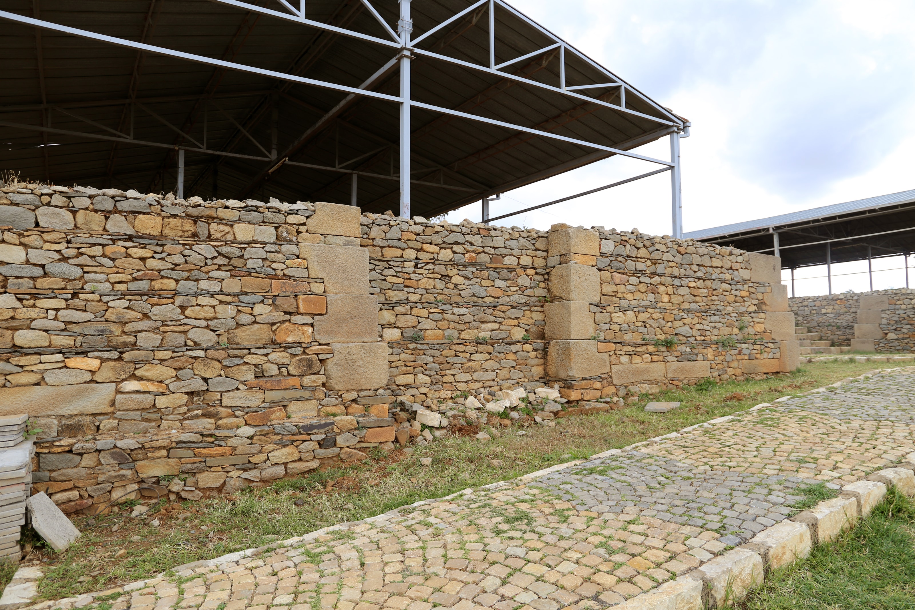 Low stone walls of dressed and rough-cut stone under a modern protective roof, beside a cobbled path, at the site called the tombs of Kaleb and Gebre Meskel