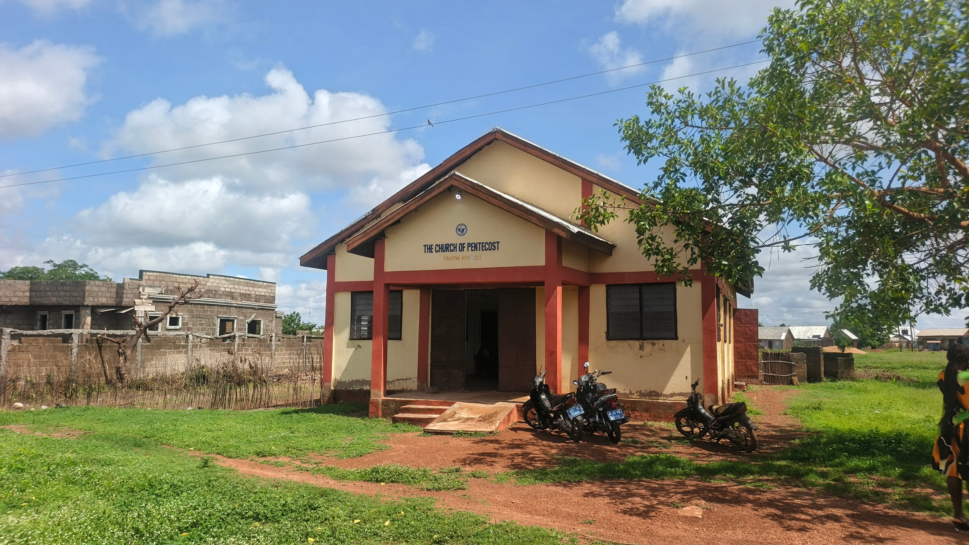 A small cream-colored church building with red trim, labeled The Church of Pentecost, standing on red earth among trees with motorcycles parked outside