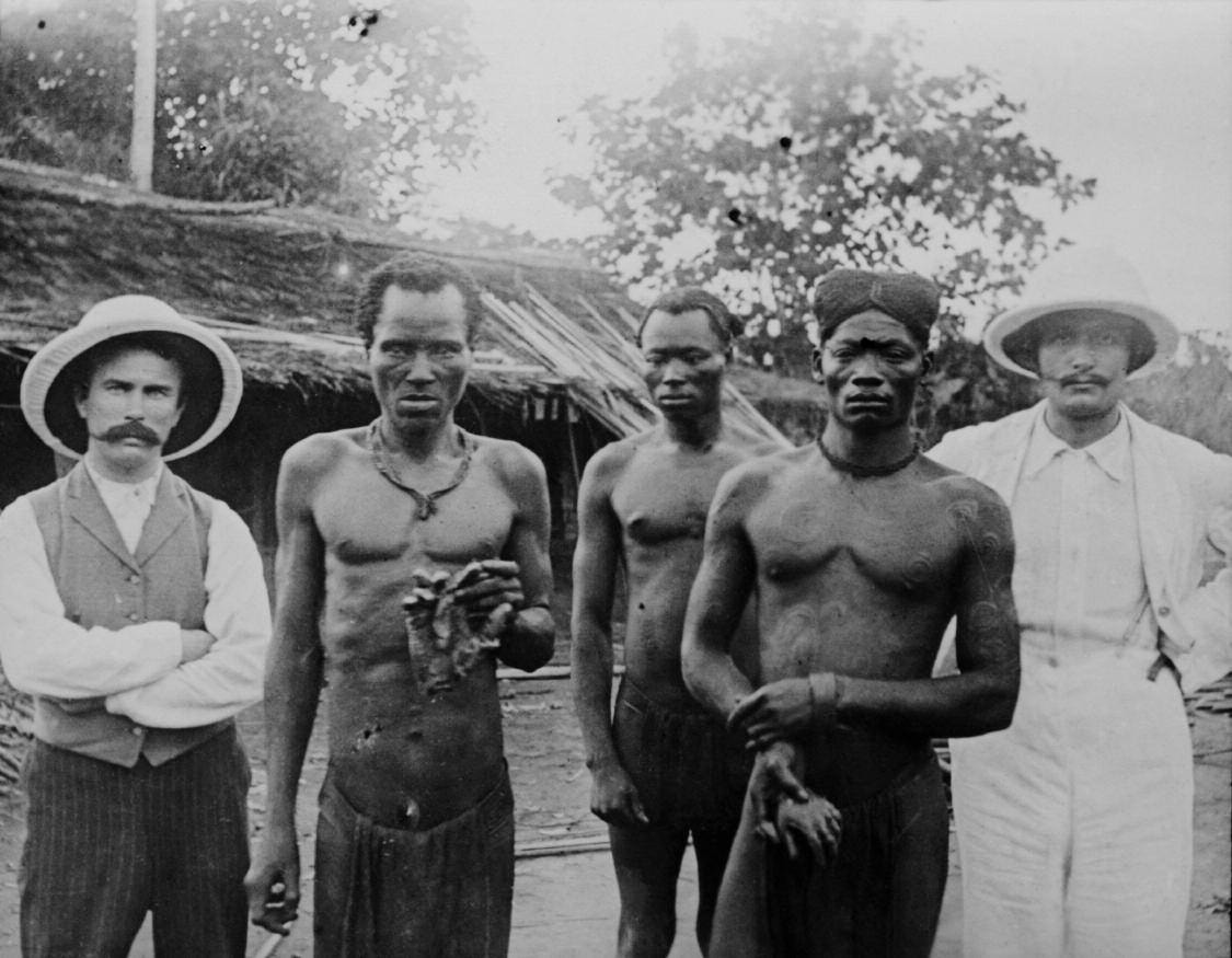 Black-and-white 1904 photograph of a group of Congolese men and two European missionaries on a veranda, with the severed hands of two victims laid before them