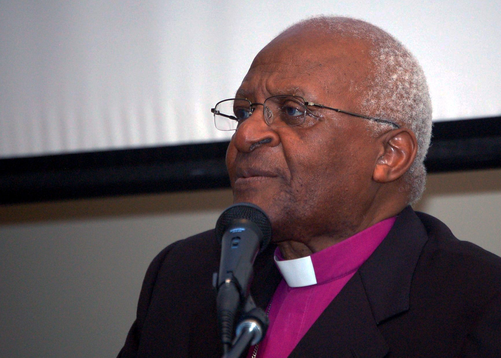 Archbishop Desmond Tutu in a purple clerical shirt and dark jacket speaking at a microphone
