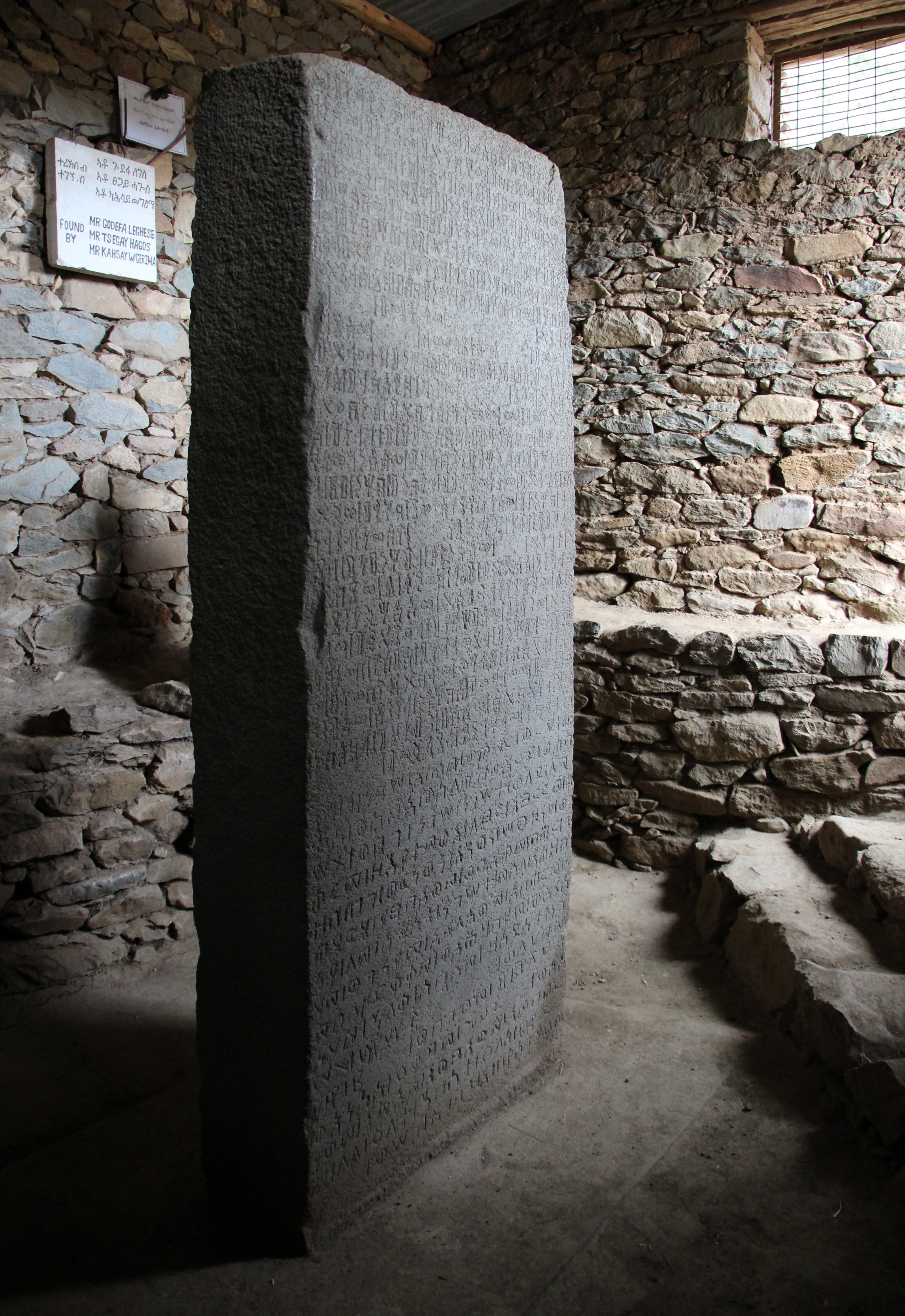 A tall dark grey stone slab covered in rows of carved script, standing inside a small stone-walled shelter