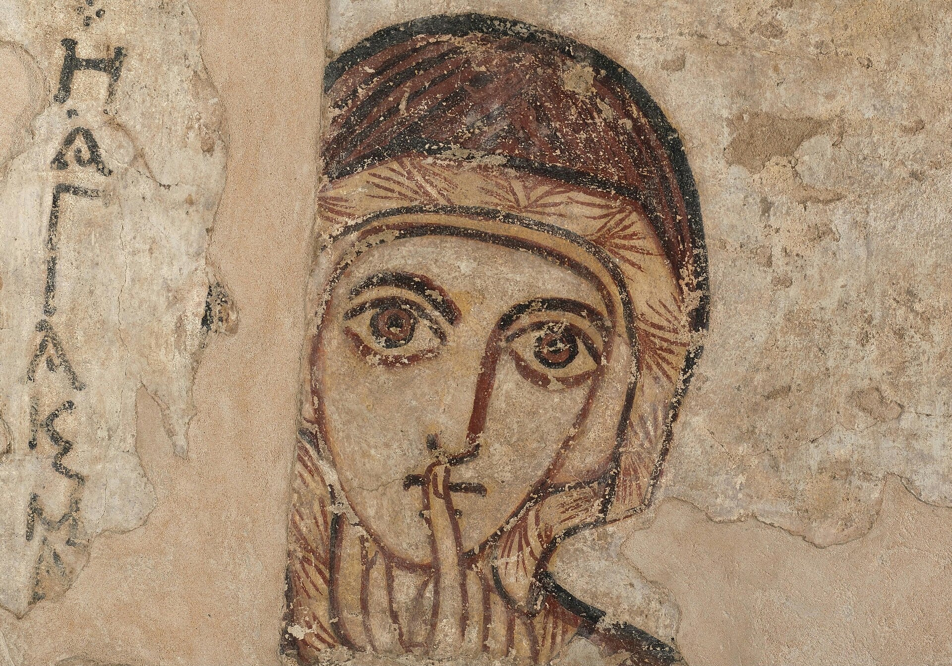 A medieval wall painting of Saint Anne, a woman in a dark head-covering, with a finger to her lips, against a pale background