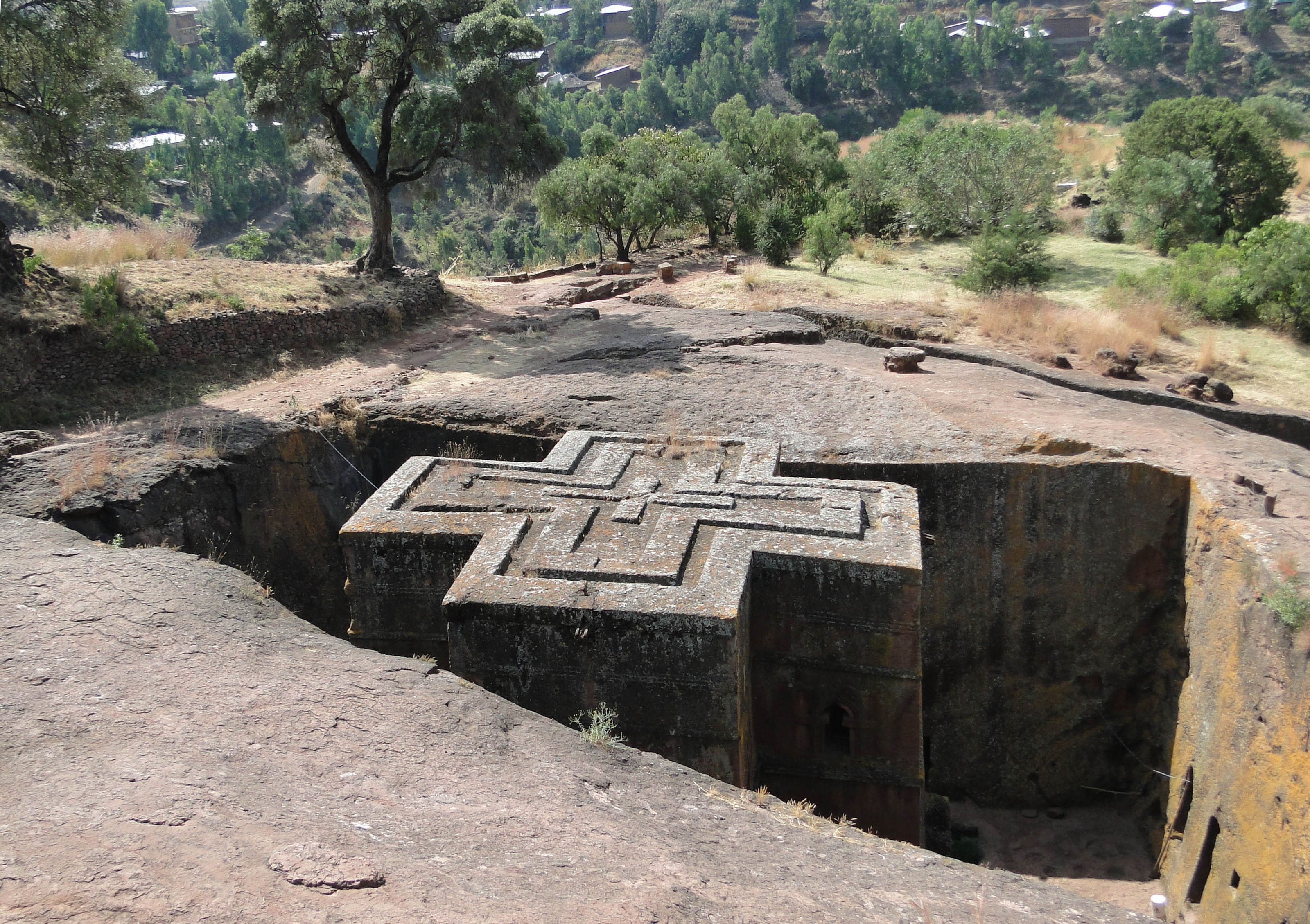A church carved in the shape of a cross, its flat roof marked with a raised cross, standing at the bottom of a deep square pit cut into reddish rock