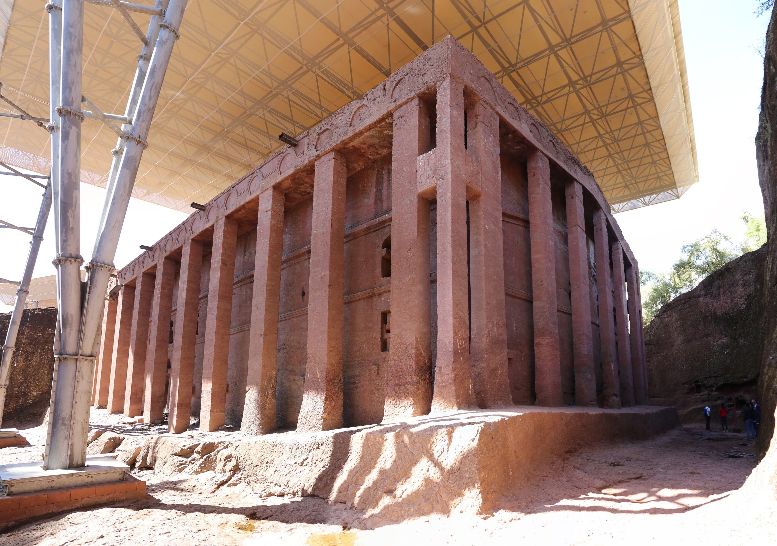 A massive rectangular church carved from red rock, surrounded by squared pillars, standing in a trench beneath a modern protective canopy