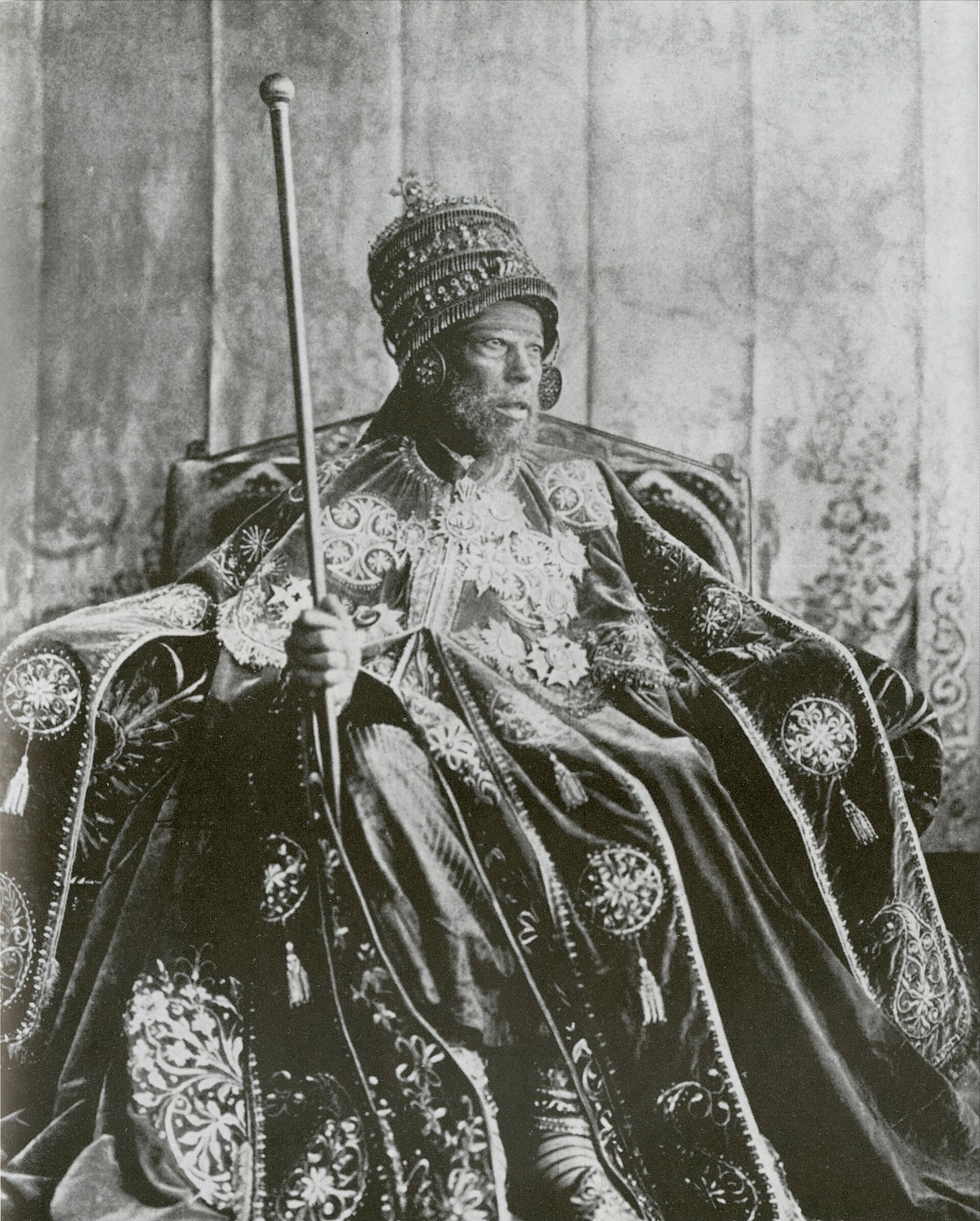 Black-and-white photograph of Emperor Menelik II seated on a throne in an embroidered coronation cape and crown, holding a tall staff