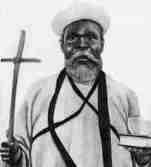 Old photograph of the Prophet William Wade Harris, a bearded man in a white robe and cap, holding a cross staff